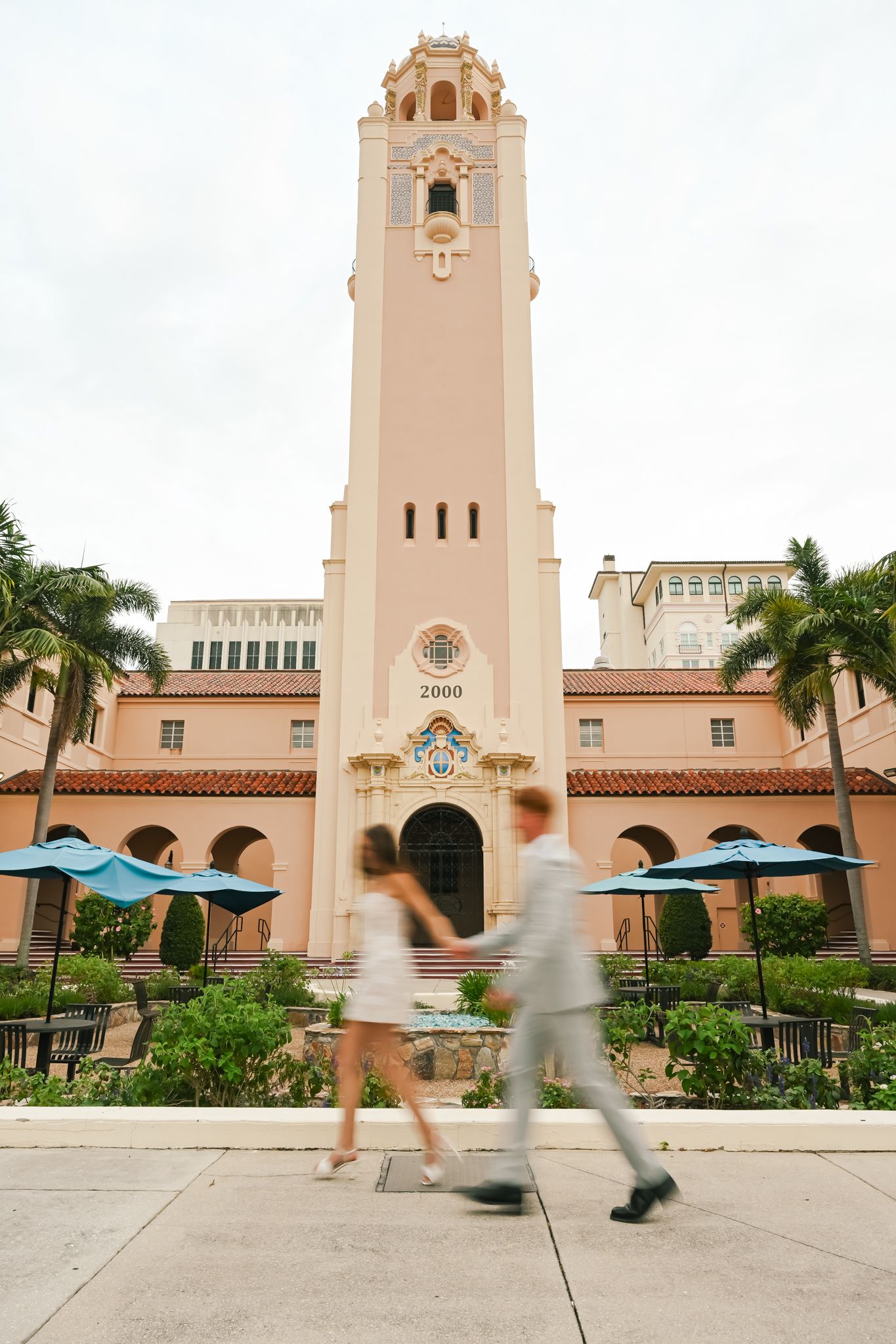 Two people walk past a pink Spanish-style building with a tall bell tower and blue umbrellas in a courtyard.