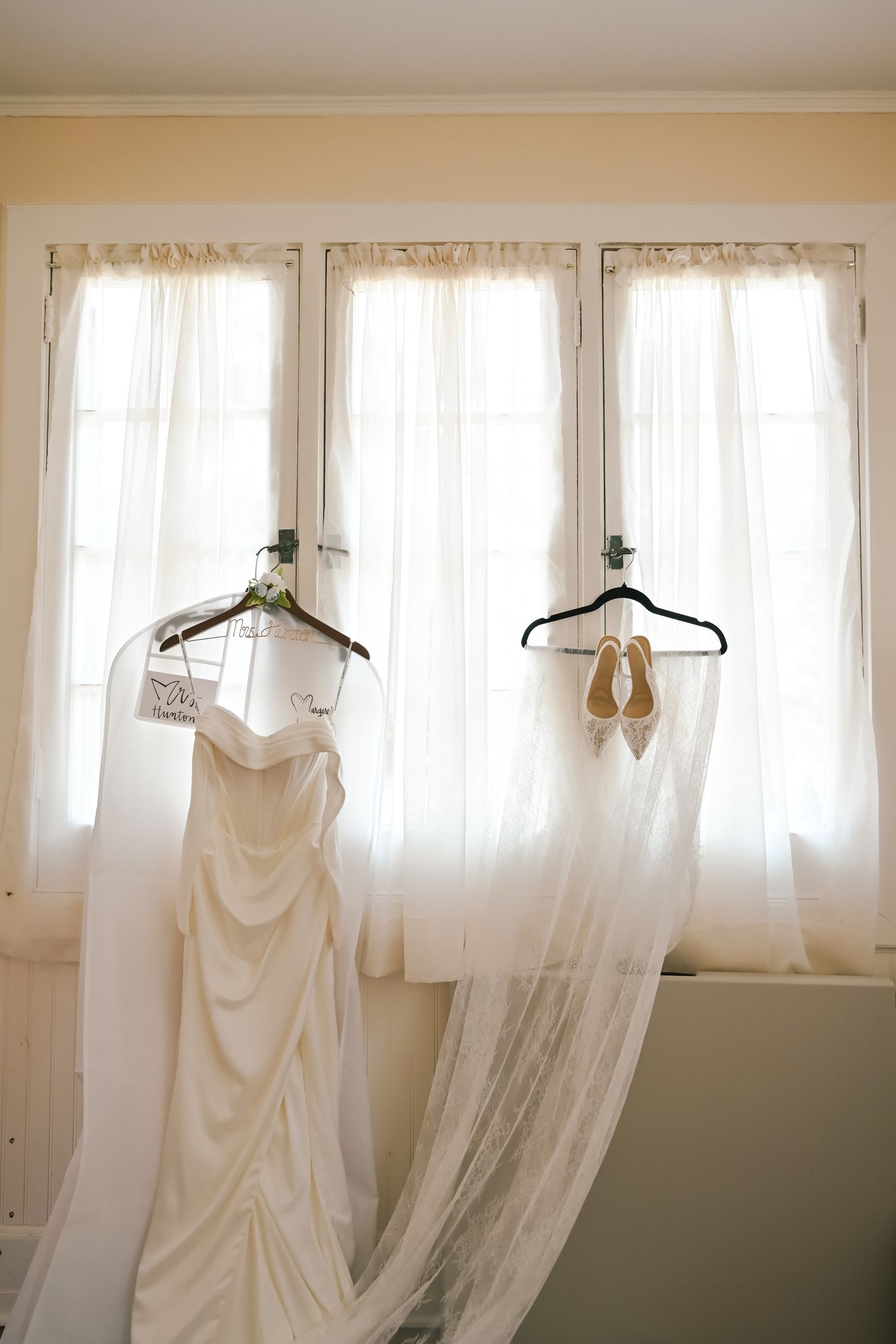 Wedding dress and veil hanging on hangers before a bright window with sheer curtains and bridal shoes displayed.