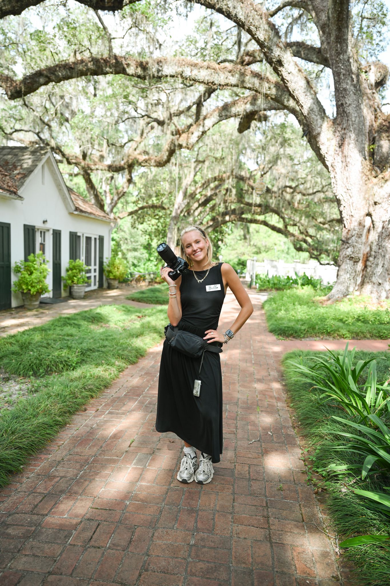 Woman in black dress holding camera on brick path under Spanish moss-draped oak trees near white building.