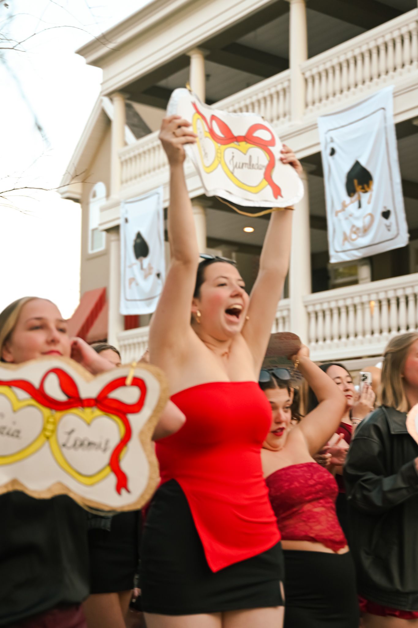 Woman in red tube top raises decorated cookie aloft at a lively outdoor sorority event with banners.