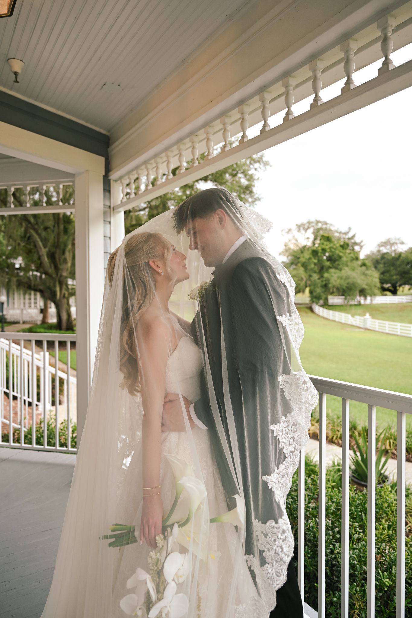 Bride and groom share an intimate moment under a lace veil on a white porch, surrounded by lush greenery.