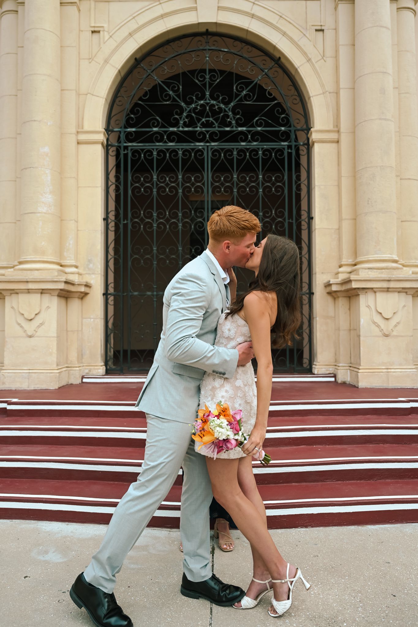 Couple kissing in front of ornate church entrance, man in gray suit, woman in floral dress holding bouquet.