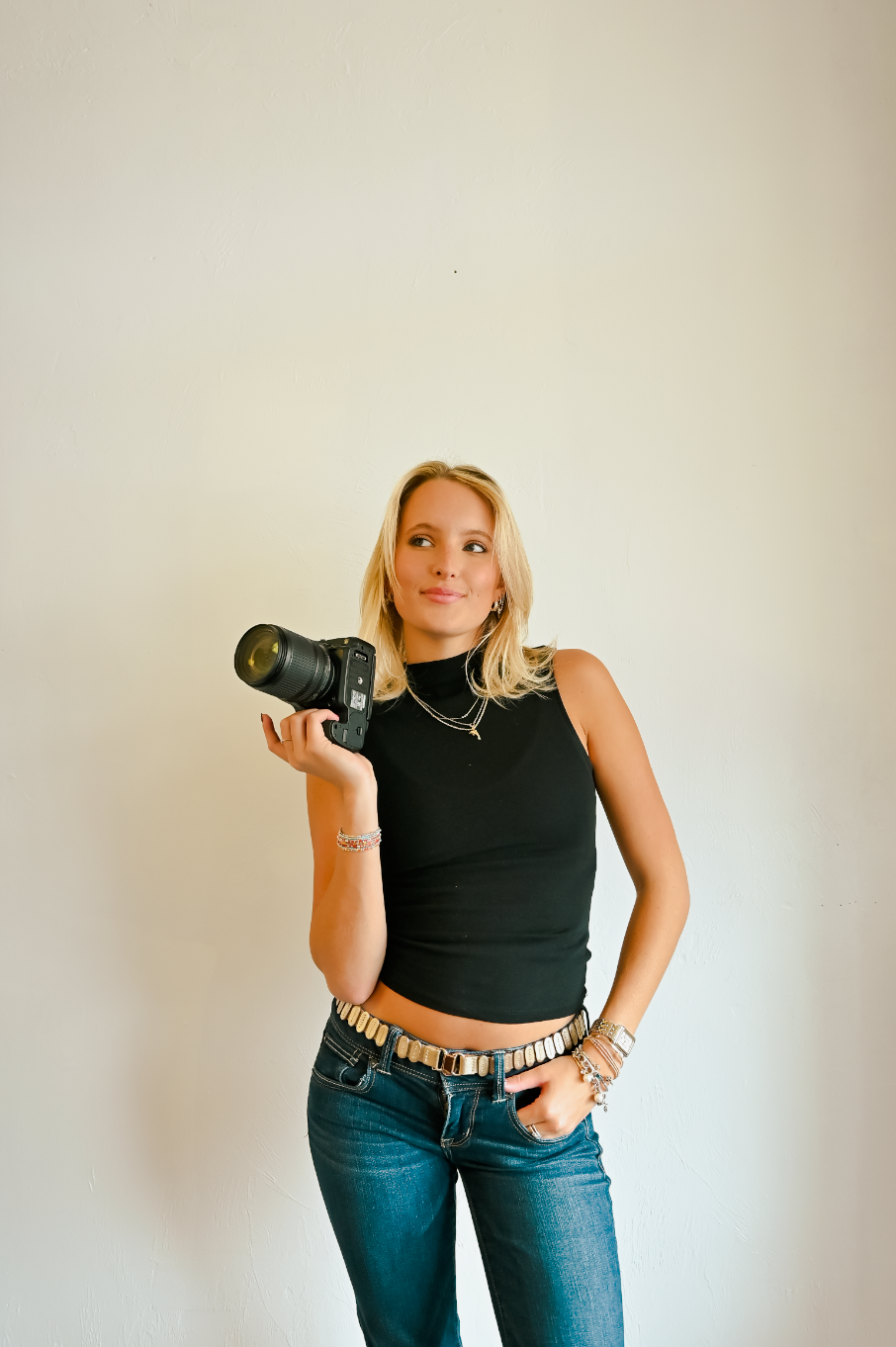 Blonde woman in black crop top and jeans holding a DSLR camera, posing against a white wall.