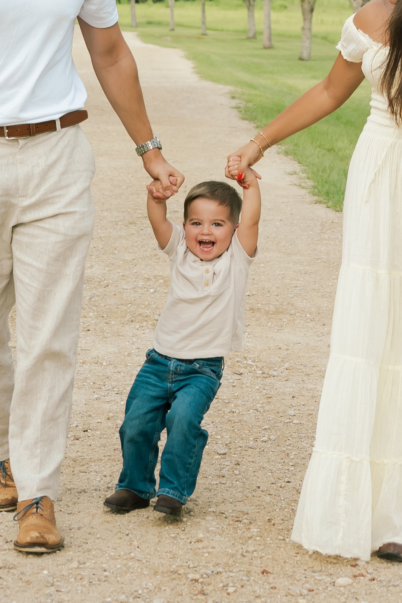 Joyful toddler boy laughing while holding hands with parents on a scenic gravel path outdoors.