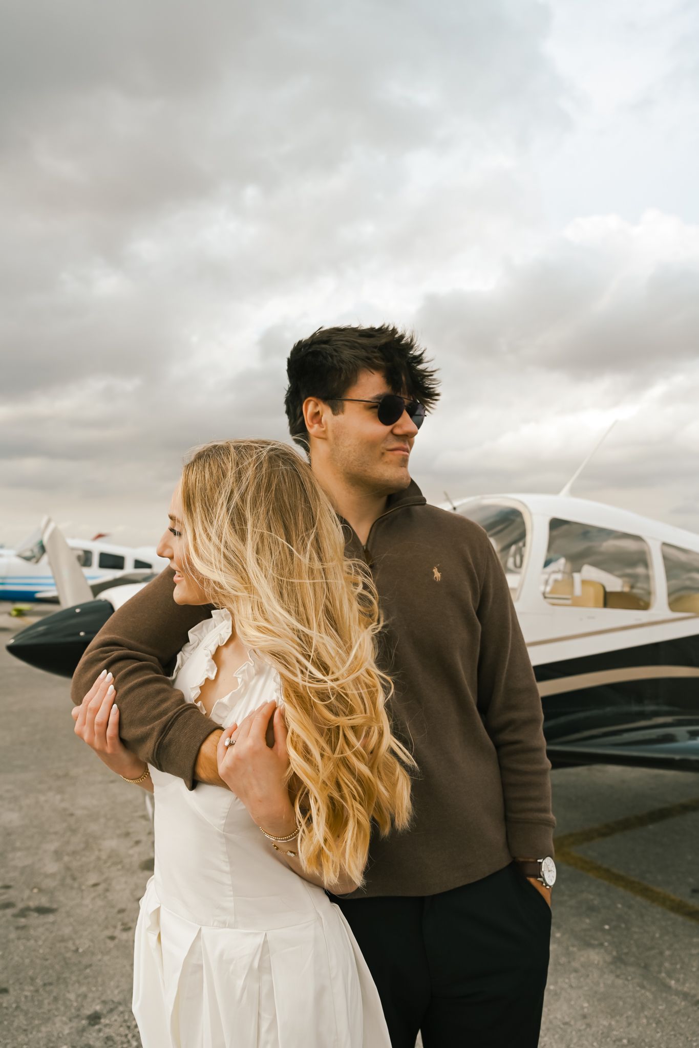 Couple embracing at an airport tarmac with a small plane behind them under a cloudy sky.