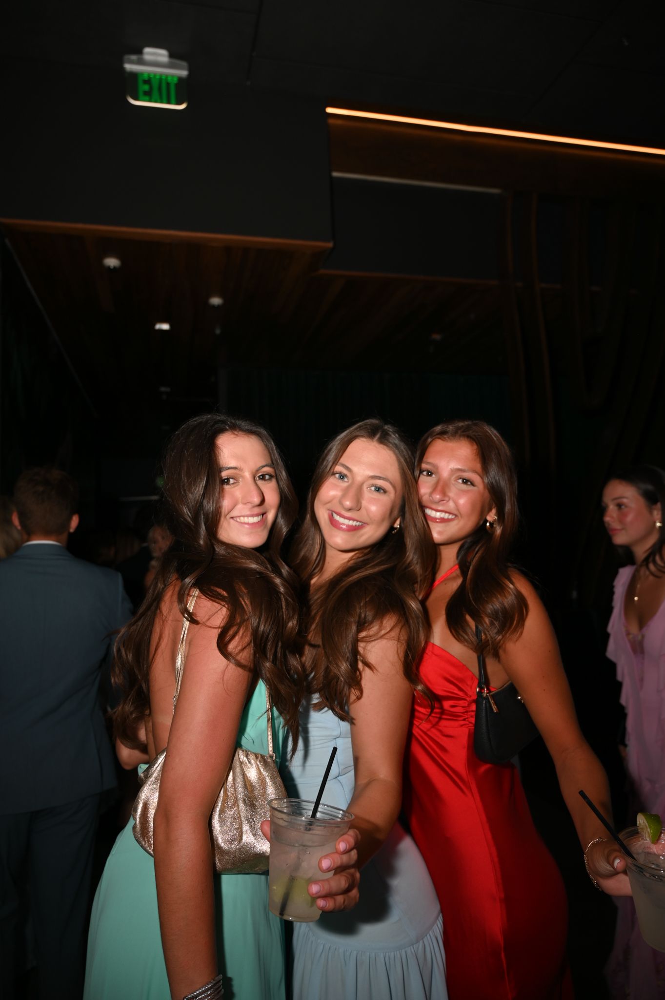 Three young women in formal dresses smiling at a party, one holding a cocktail drink.