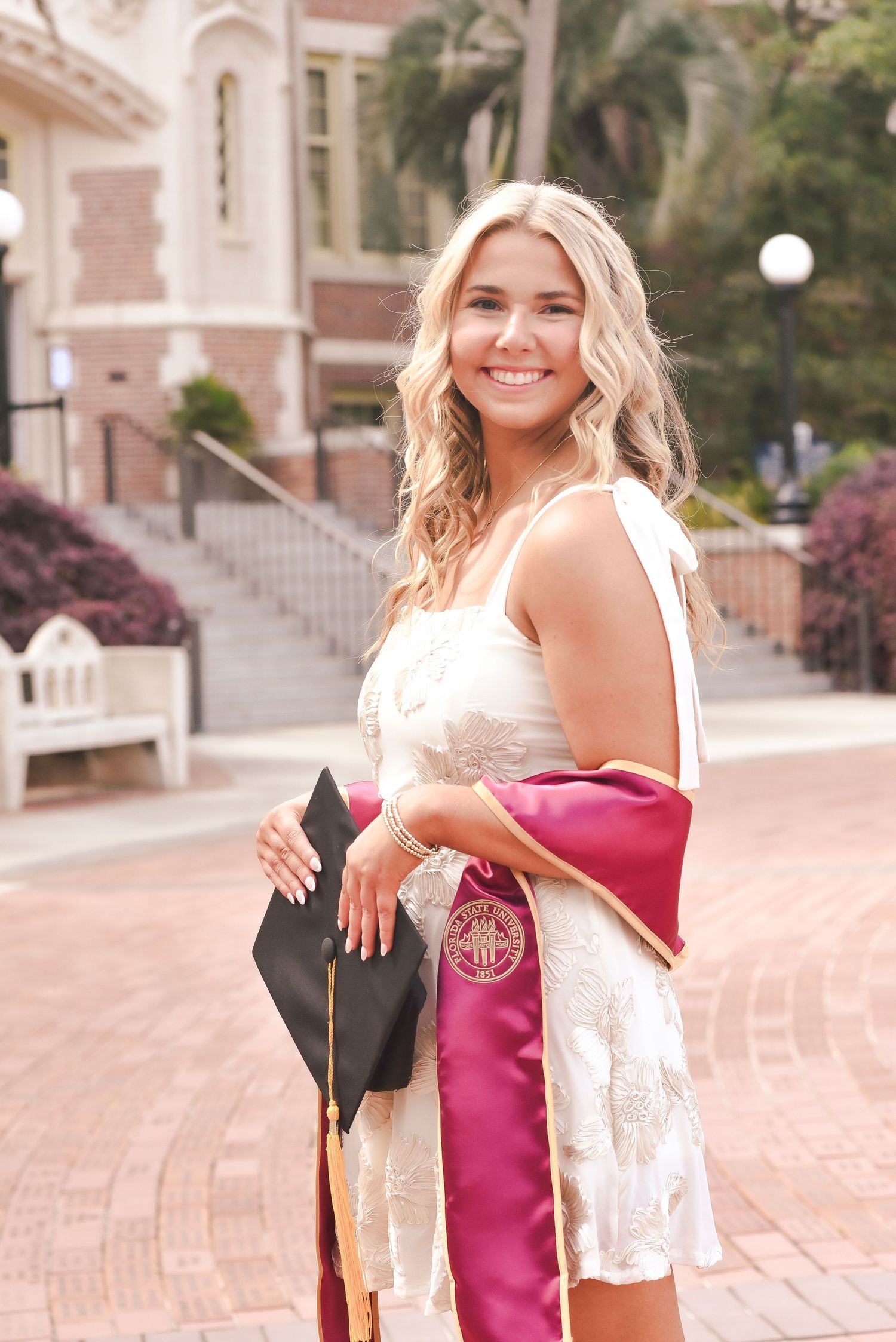 Graduate in white lace dress holds cap and graduation stole on campus walkway.