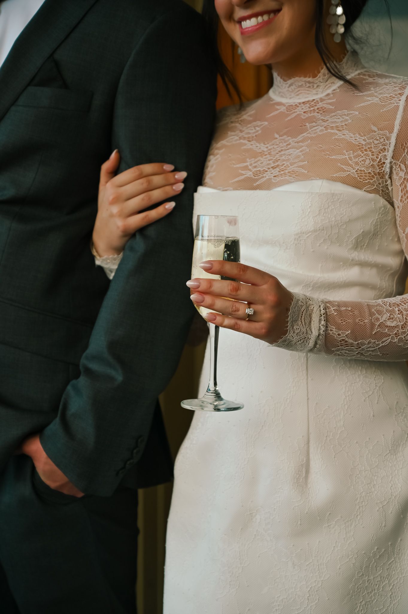 Bride in lace wedding dress holding champagne flute, smiling beside groom in dark suit at wedding reception.