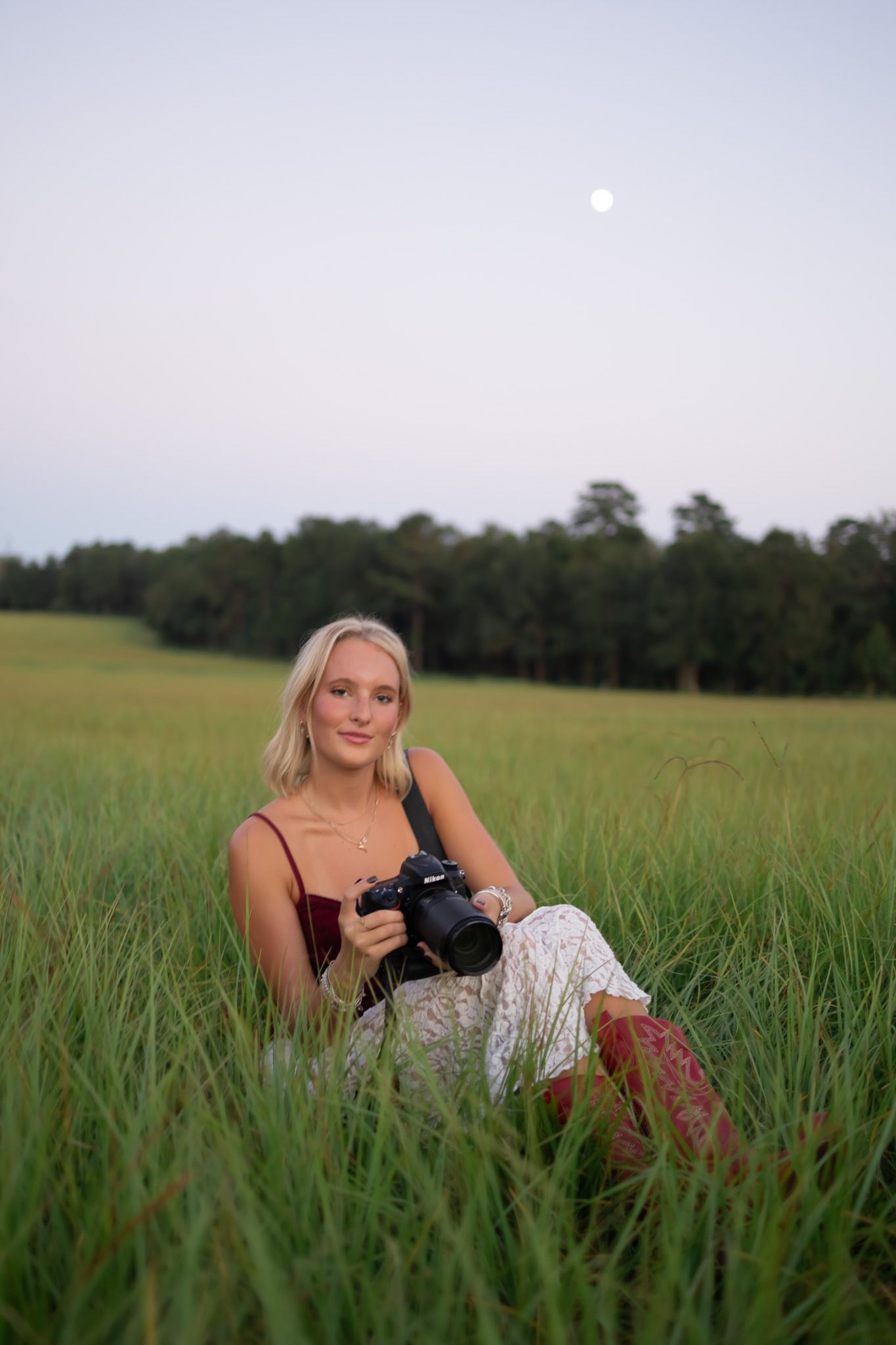 Young blonde woman sitting in tall green field holding a camera with full moon and trees in background.
