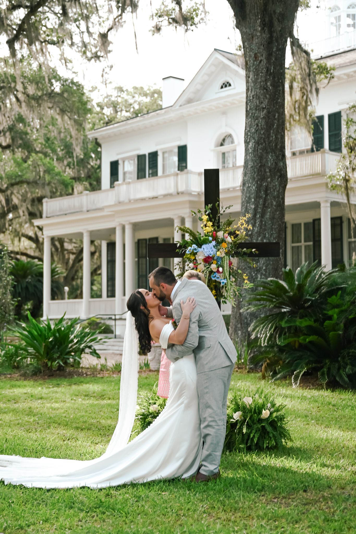 Bride and groom share first kiss outdoors at elegant white Southern plantation wedding venue surrounded by oak trees.