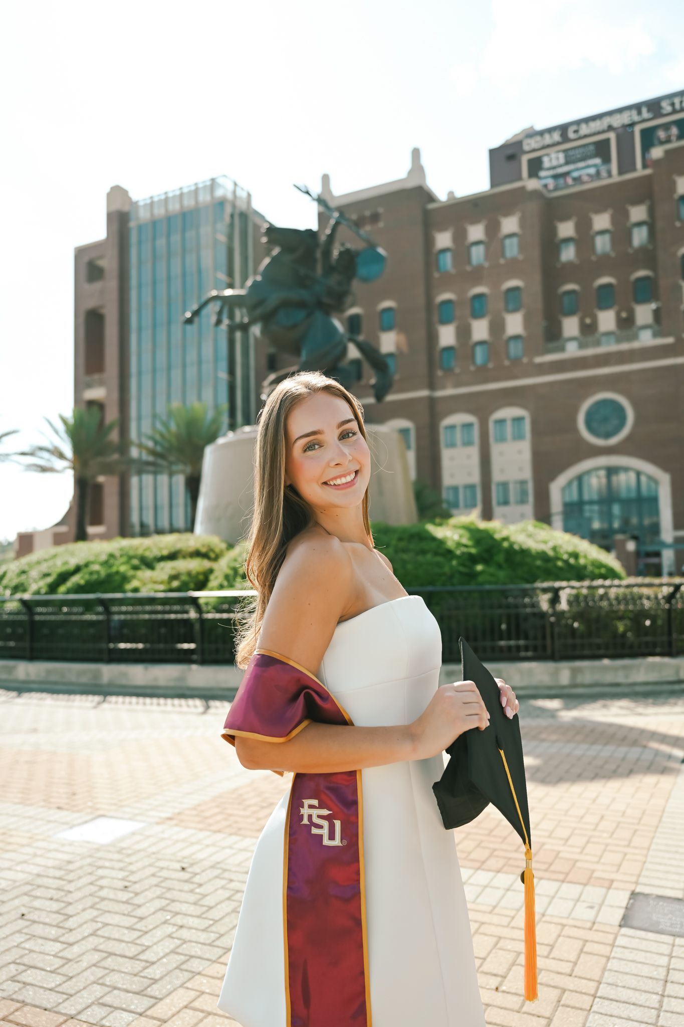 FSU graduate smiles in white dress, maroon sash, and cap outside Doak Campbell Stadium in Tallahassee, FL.
