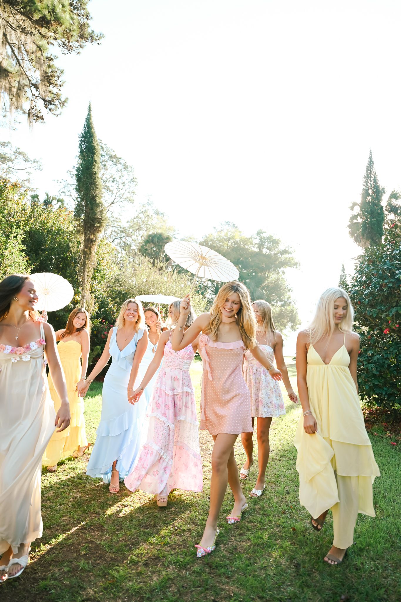 Group of women in pastel dresses walking outdoors in a sunny garden, some holding white parasols.