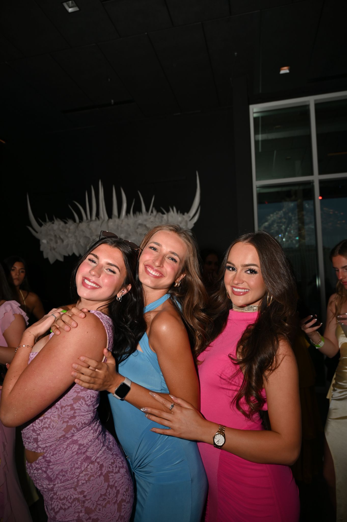 Three young women in colorful formal dresses smile and pose together at a prom or formal event.