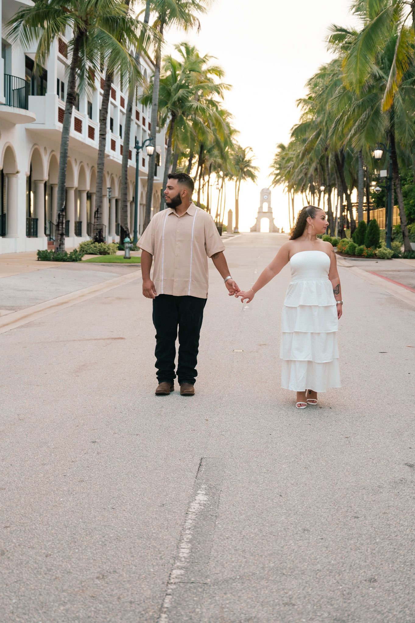 Couple holding hands on a palm-lined street, man in beige shirt and black pants, woman in white tiered dress, at sunset.