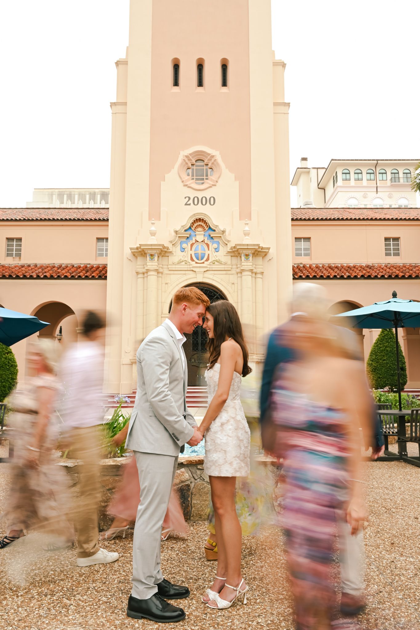 Couple kisses holding hands in front of a pink building dated 2000, with blurred guests passing by.