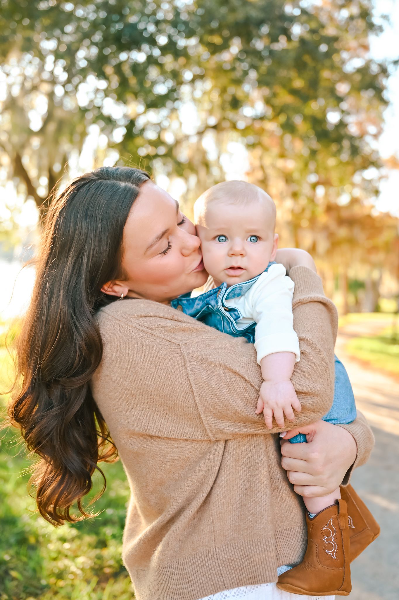 A person in a tan sweater gives loving kisses to a baby in blue outdoors.