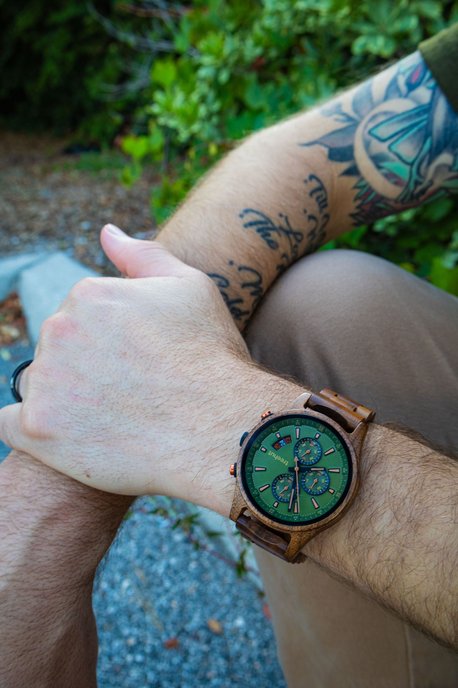 Tattooed arm wearing a wooden watch with green chronograph dial.