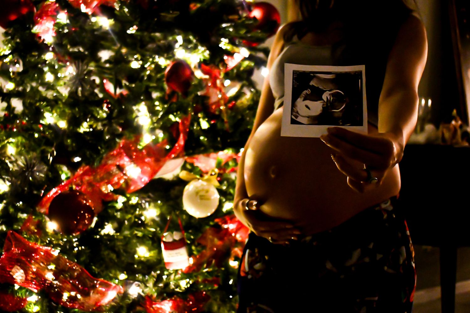 Pregnant woman holding ultrasound photo in front of illuminated Christmas tree with red ornaments and lights.