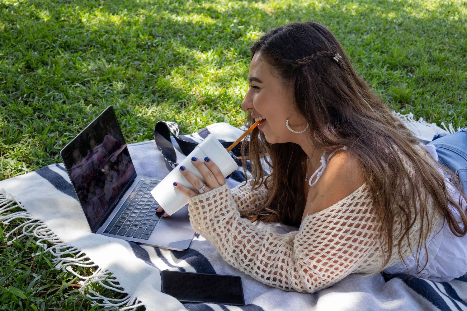 Person lying on blanket in sunny grass with laptop and camera.