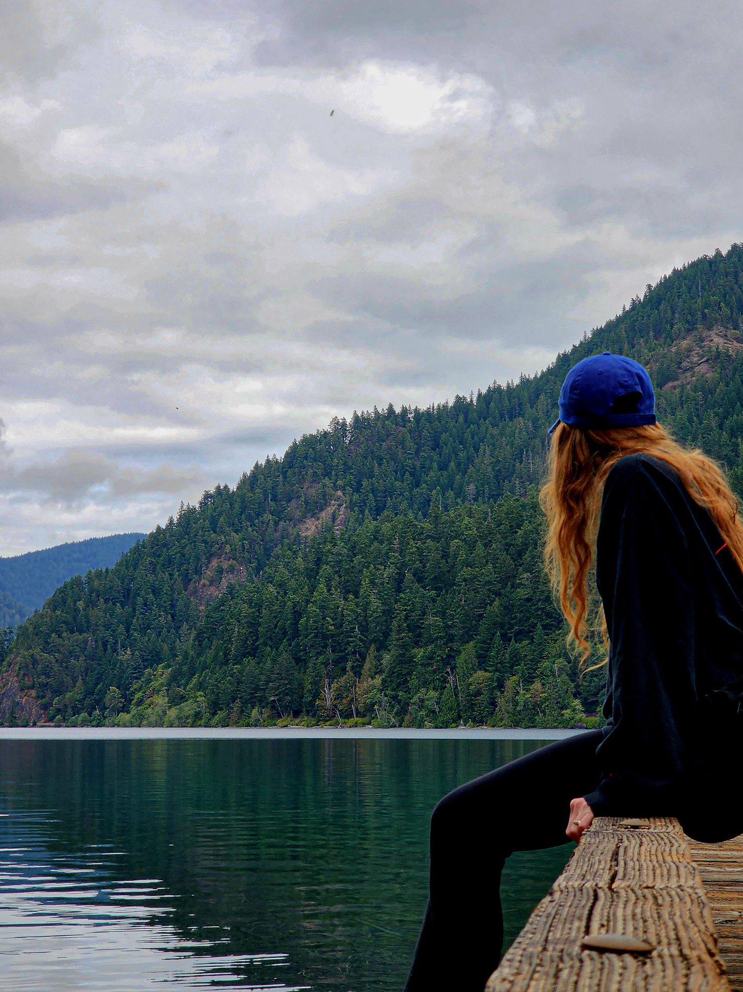 A serene lake view with evergreen-covered mountains and cloudy skies reflected in calm waters from a wooden dock.