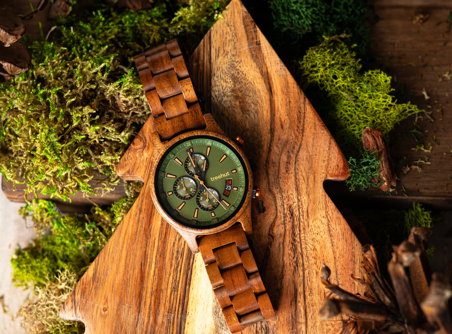 A wooden watch with green face rests on rustic wood surrounded by moss.