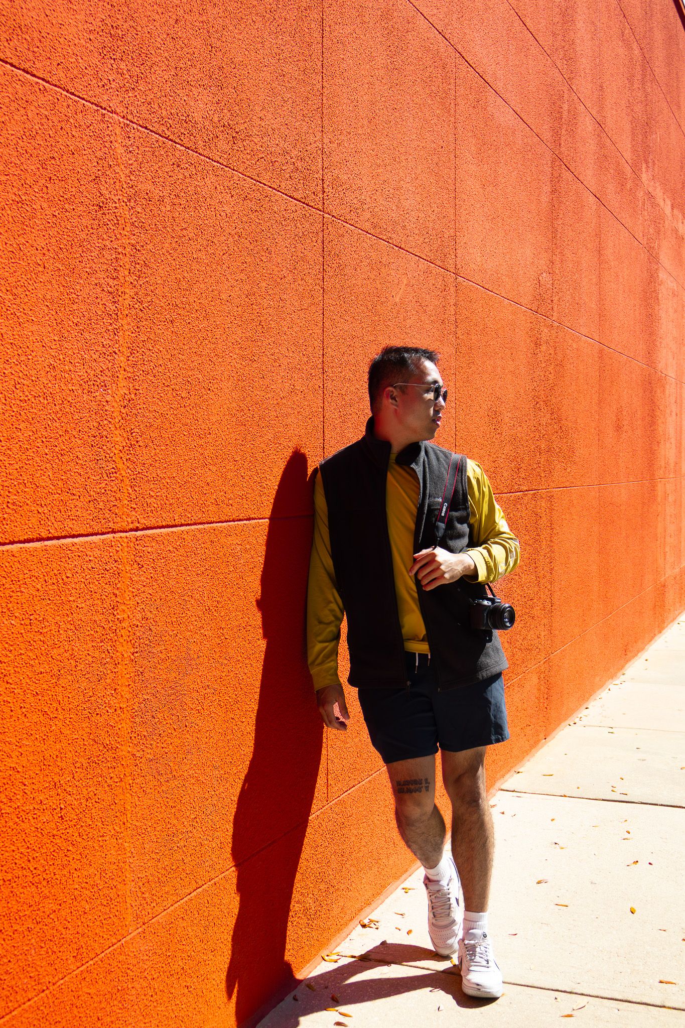 Person in yellow shirt and black vest crouches against vibrant orange wall casting dramatic diagonal shadow.