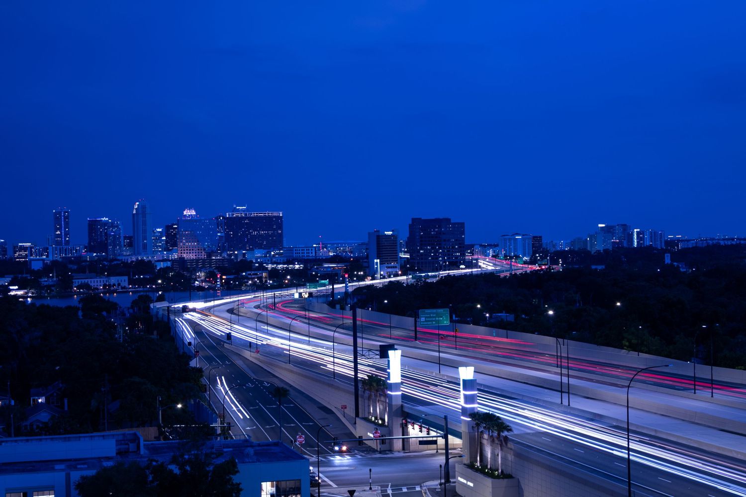 Long exposure night shot of light trails on highway with skyline illuminated against dark blue sky.