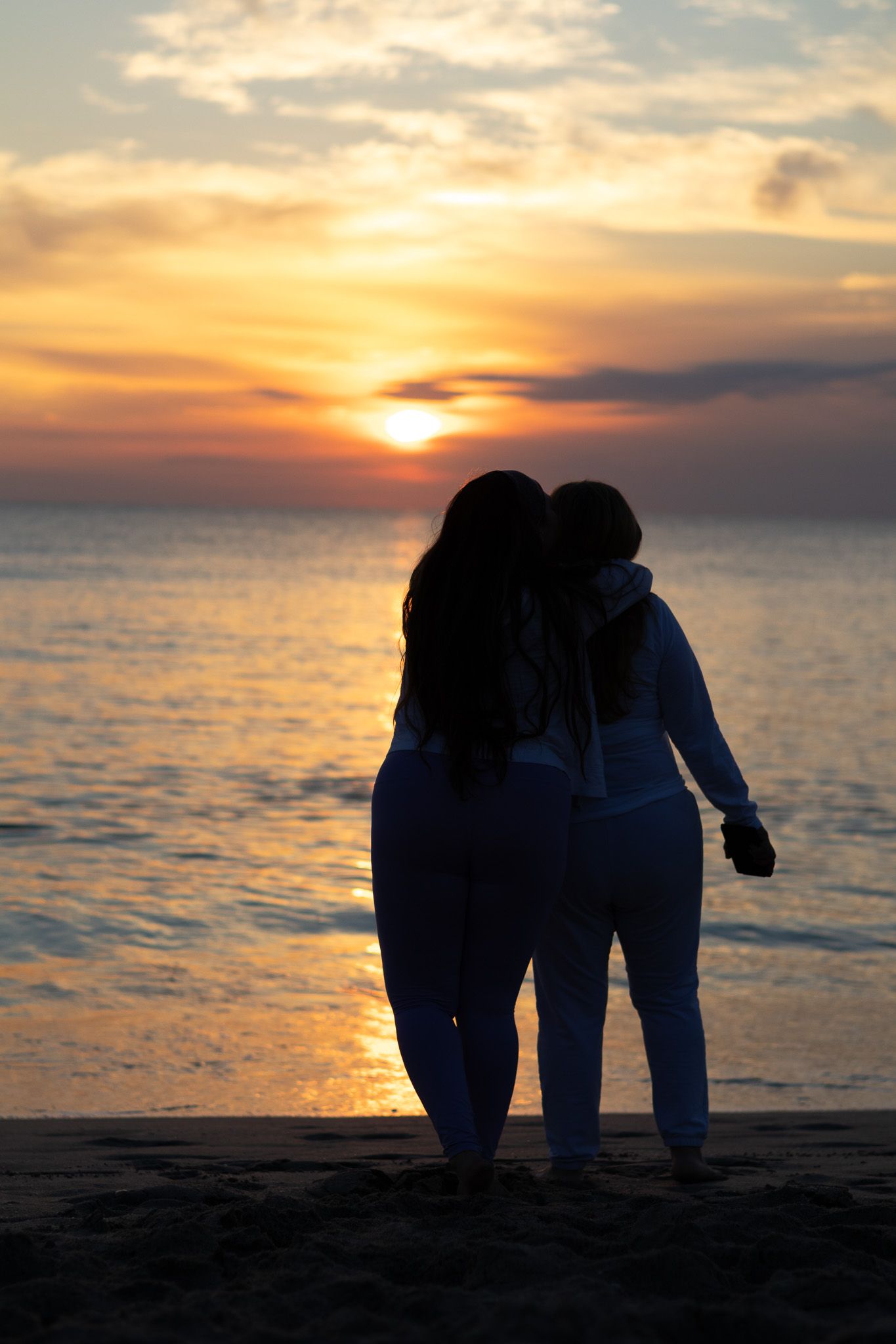 Silhouetted figures stand together watching a vibrant orange sunset over calm ocean waters.