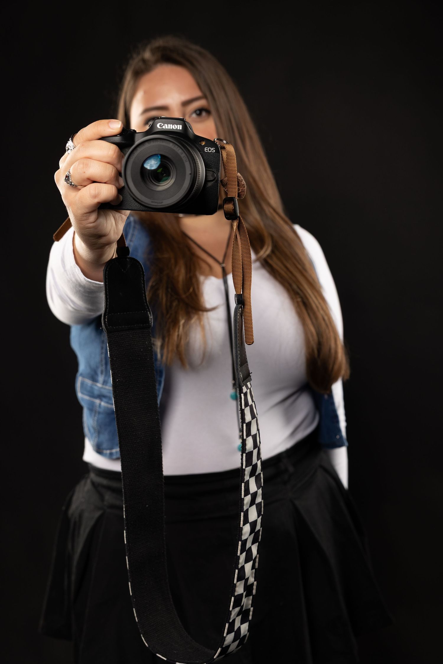 A photographer aims a professional DSLR camera while wearing casual attire against a black backdrop.