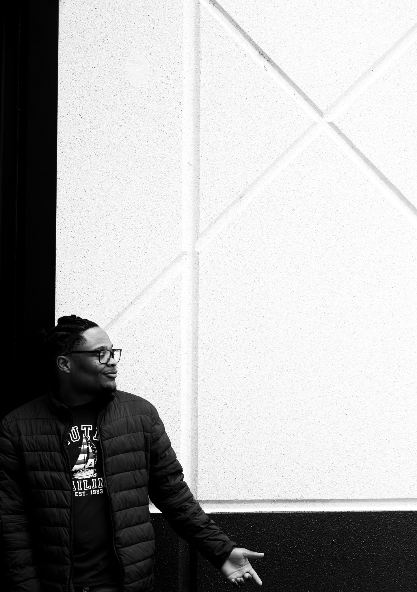 Person in winter coat and glasses sitting against minimalist white and black wall.