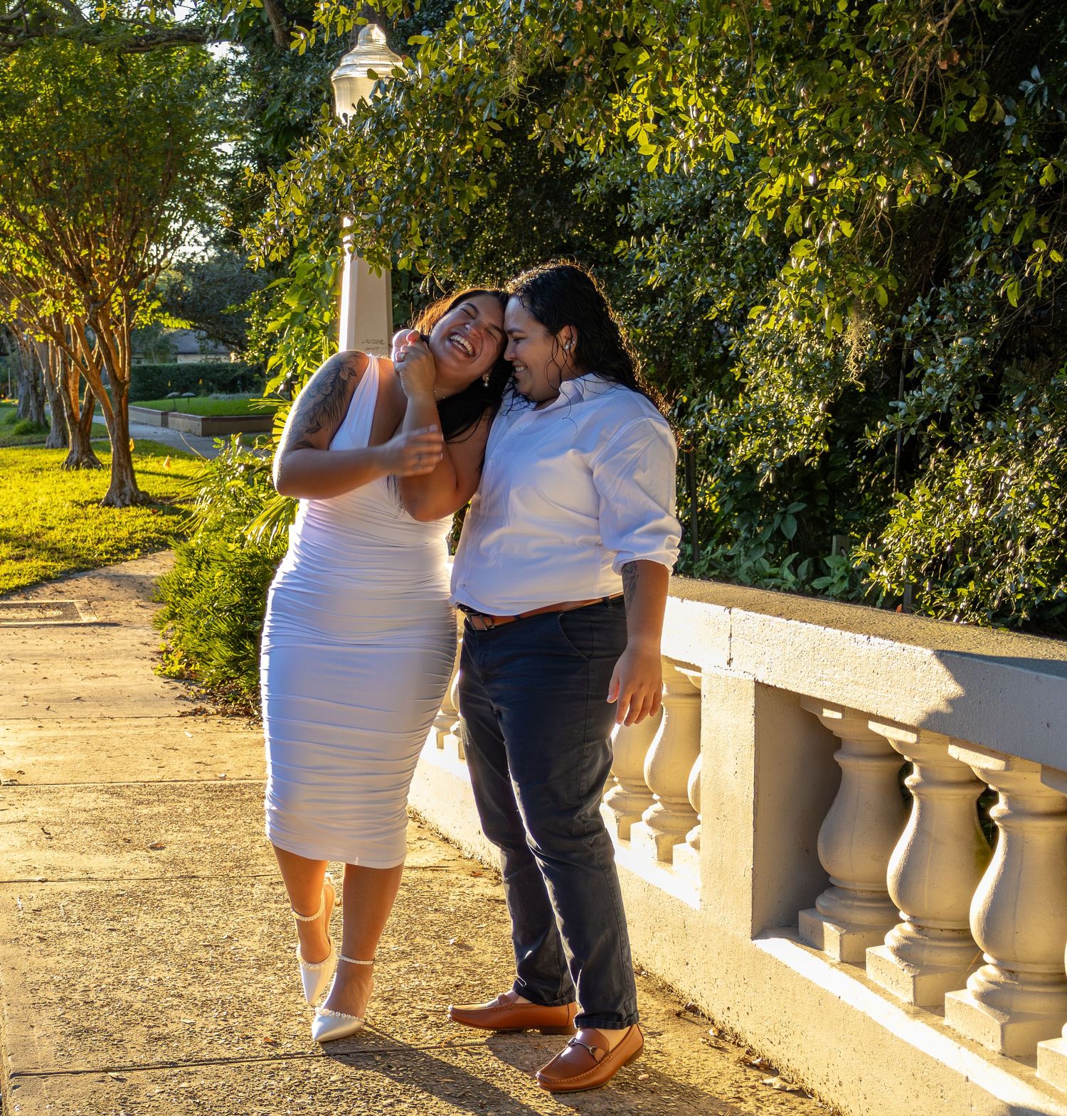 A couple shares an intimate moment along a sunlit garden path with white stone balustrades and lush greenery.
