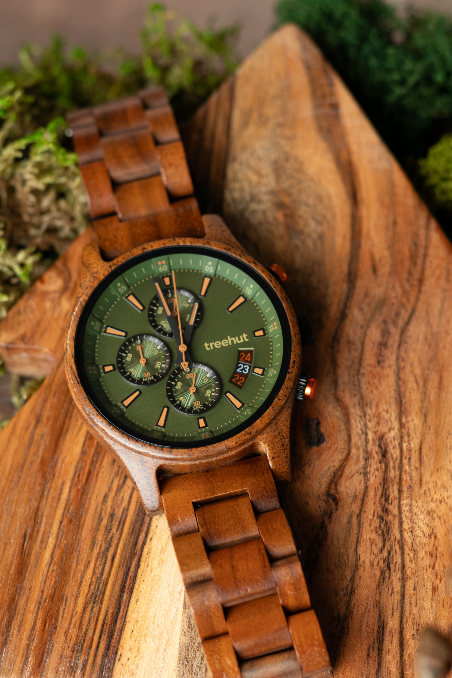 A wooden chronograph watch with green dial displayed on rustic wood with moss accents.