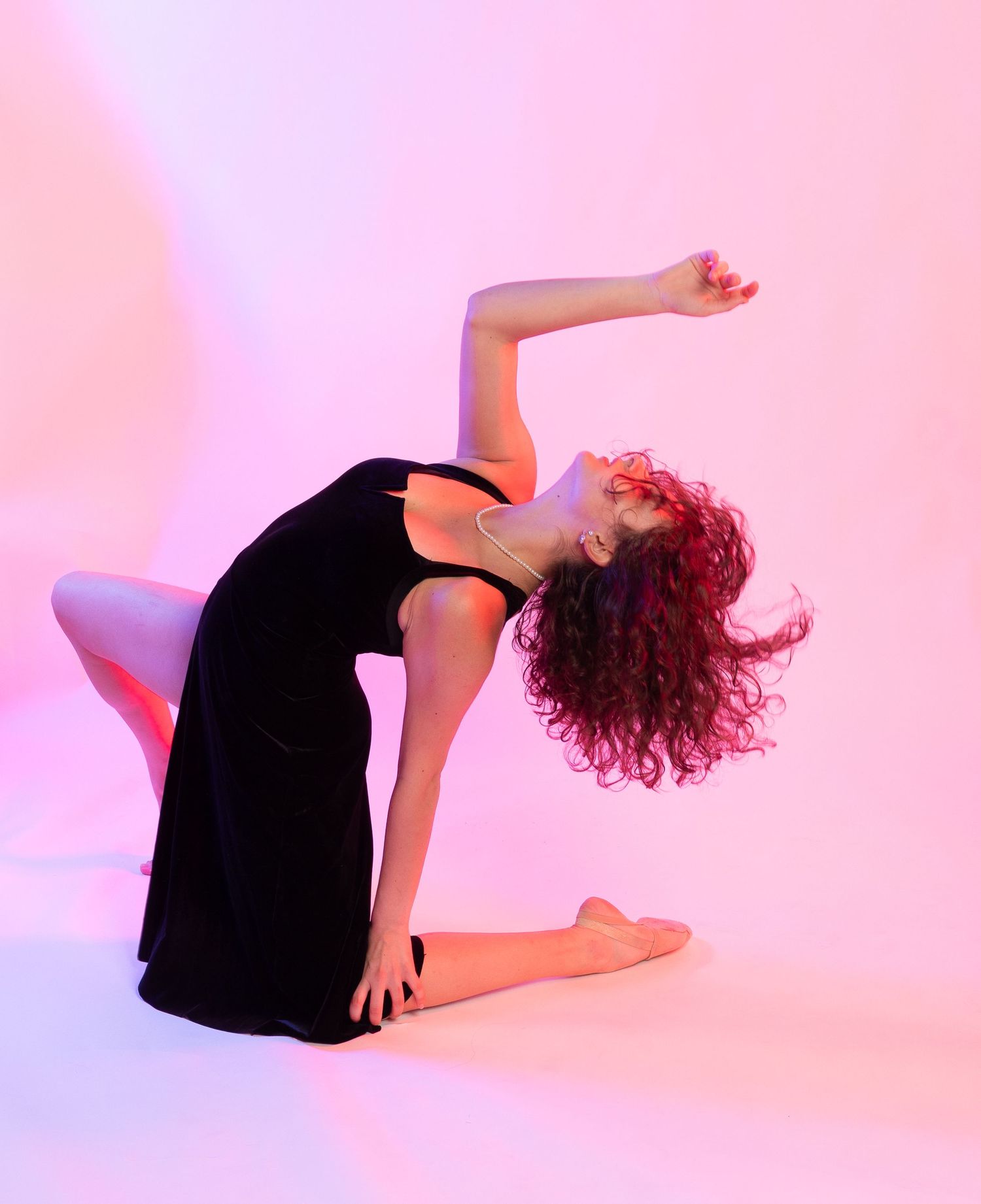 Dancer in black dress performs graceful backbend pose against pink backdrop with curly hair flowing.
