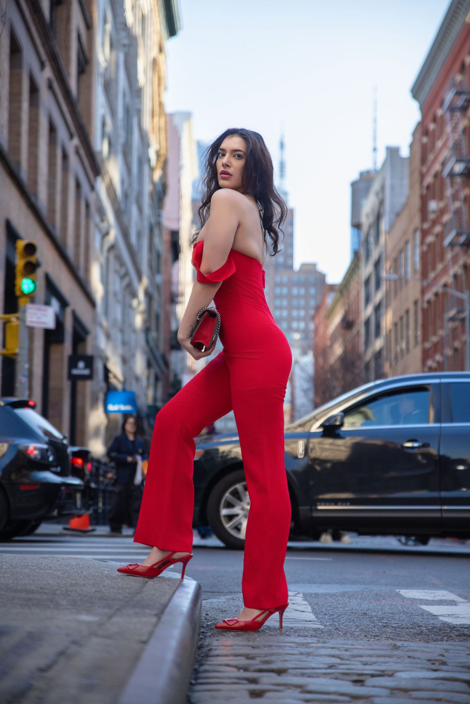 Woman in red off-shoulder jumpsuit and heels posing on urban city street with buildings in background.