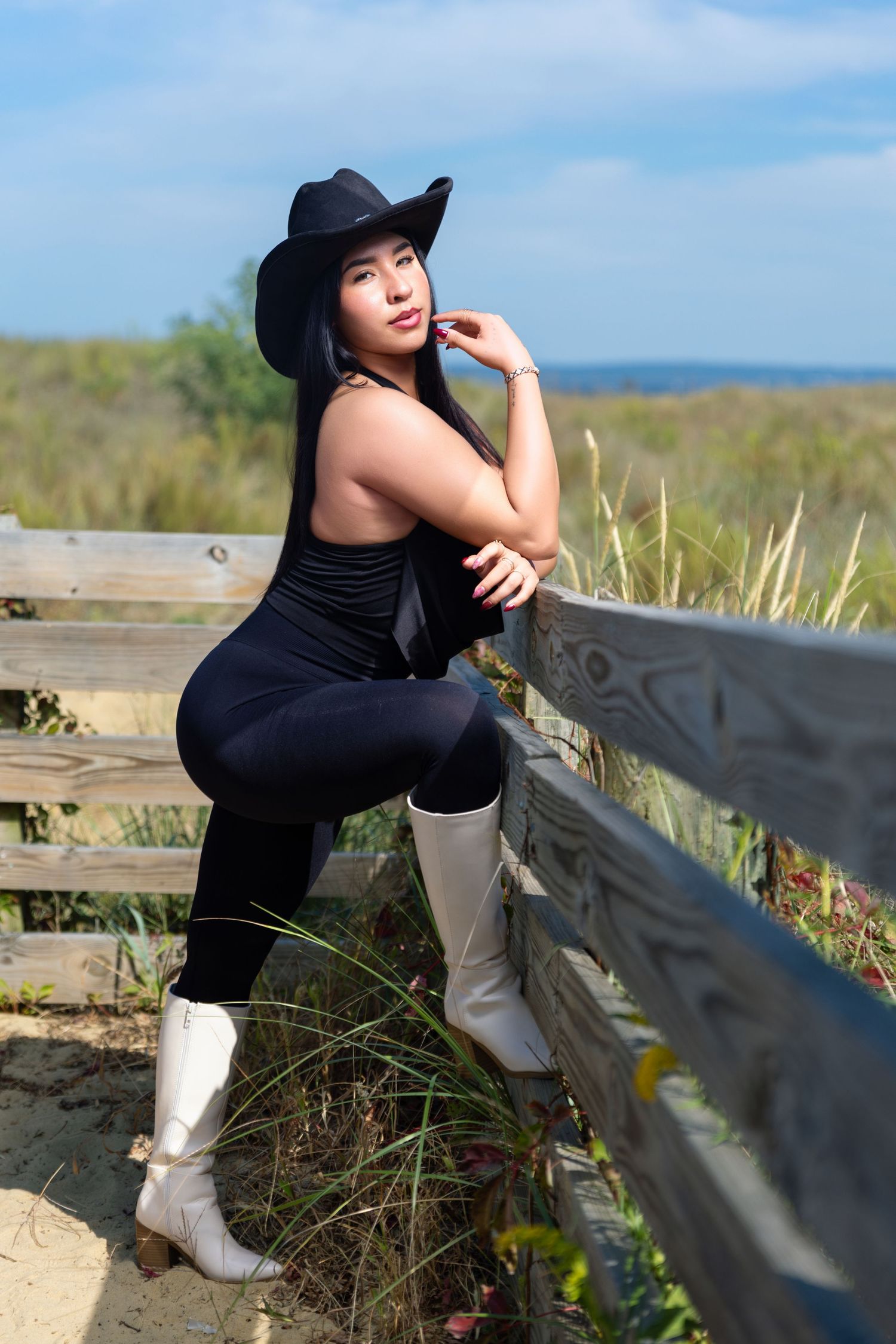 Asian woman in black outfit and cowboy hat posing by wooden fence in nature.