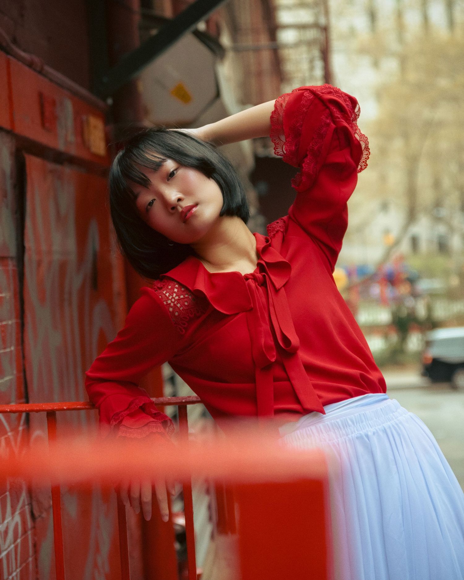 Young woman in red ruffled blouse and white skirt posing on red fire escape in urban setting.