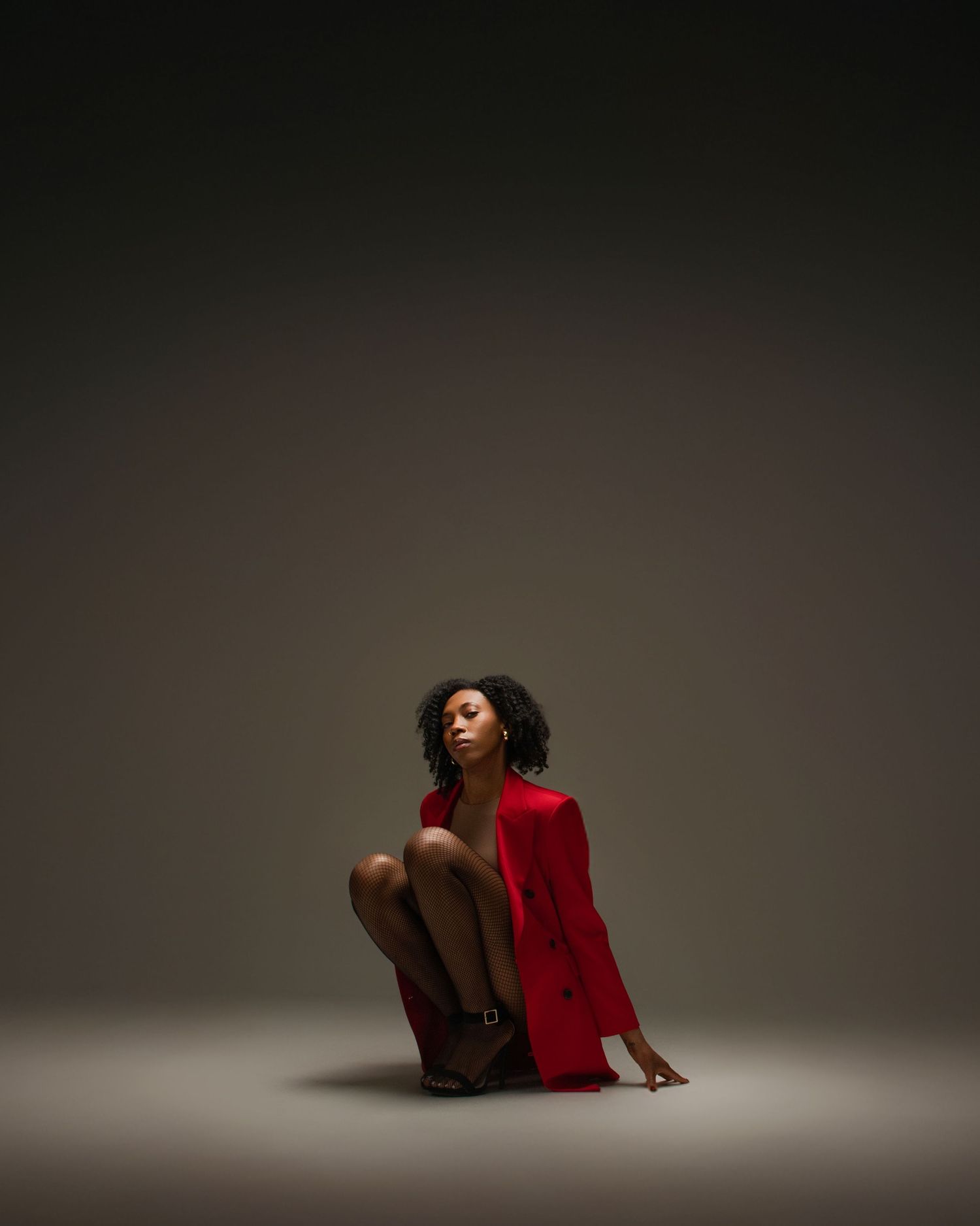 Black woman in red blazer sitting gracefully on studio floor with dramatic moody lighting.