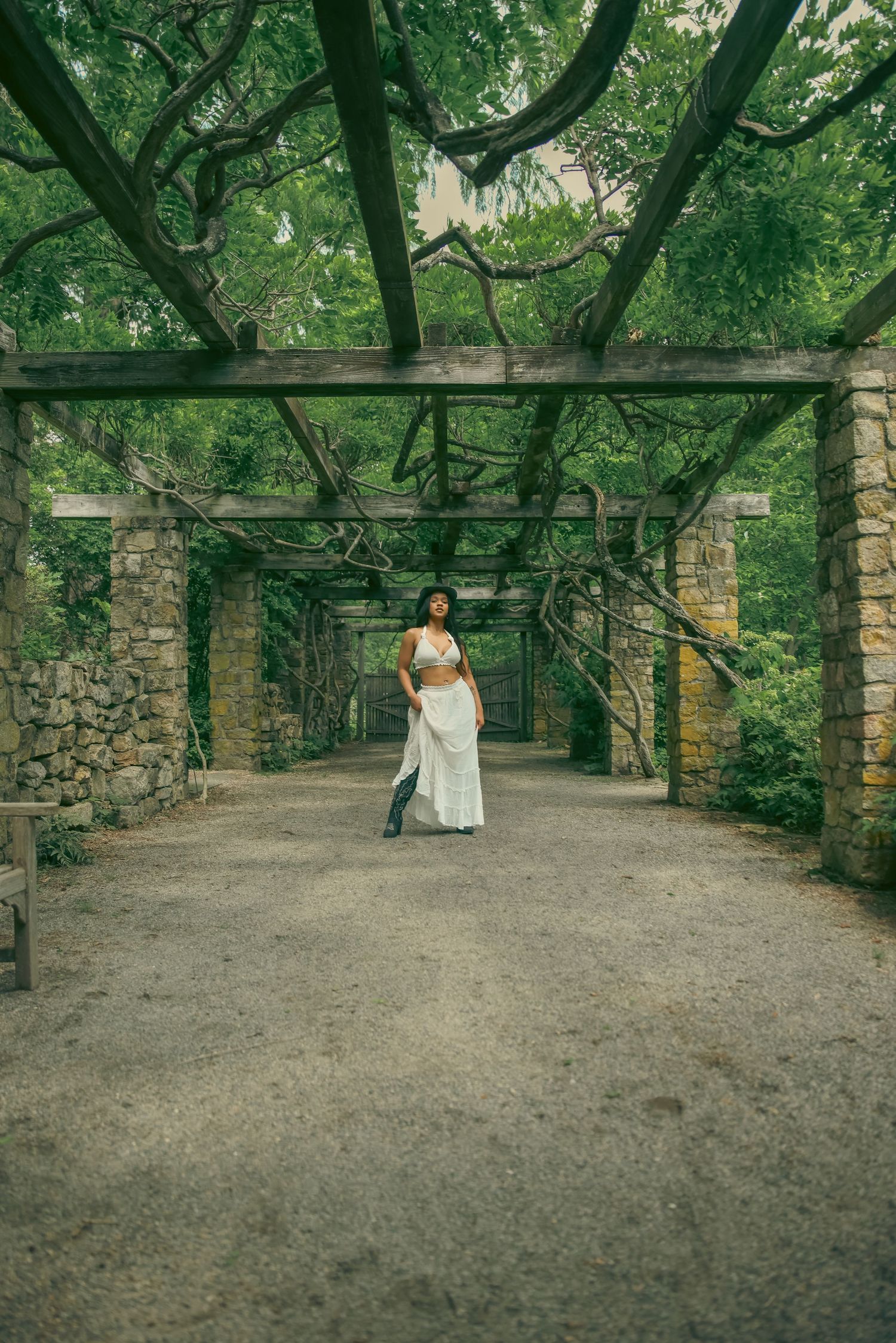 Woman in white crop top and flowing skirt walking through a rustic stone pergola tunnel surrounded by greenery.
