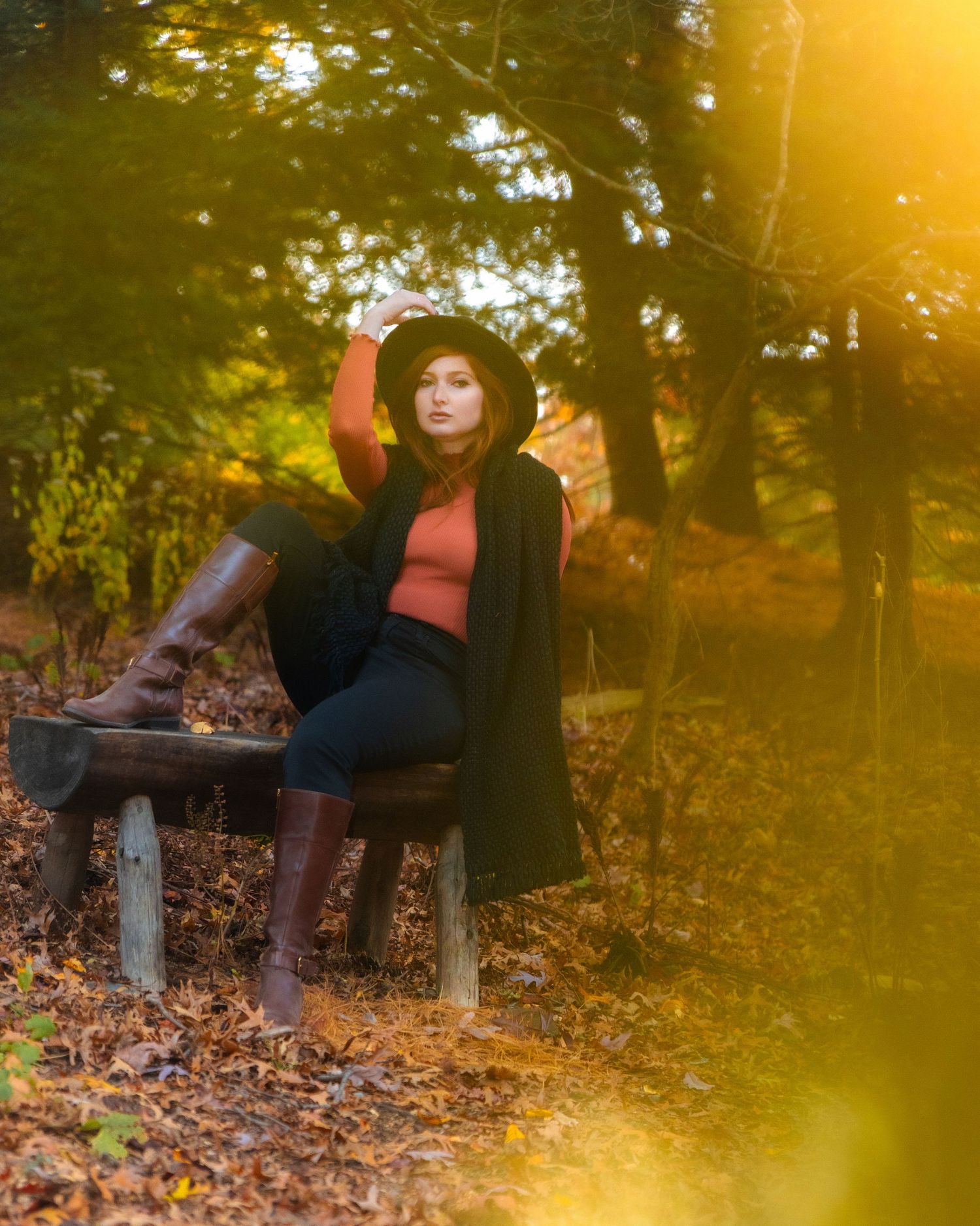 Woman in black coat, rust top, and brown boots sitting on a bench in a golden autumn forest.