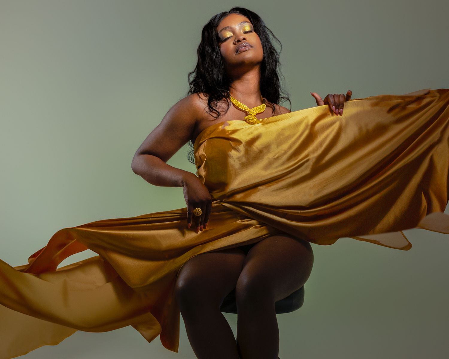 Elegant woman in flowing golden satin dress posing gracefully against a soft green studio background.