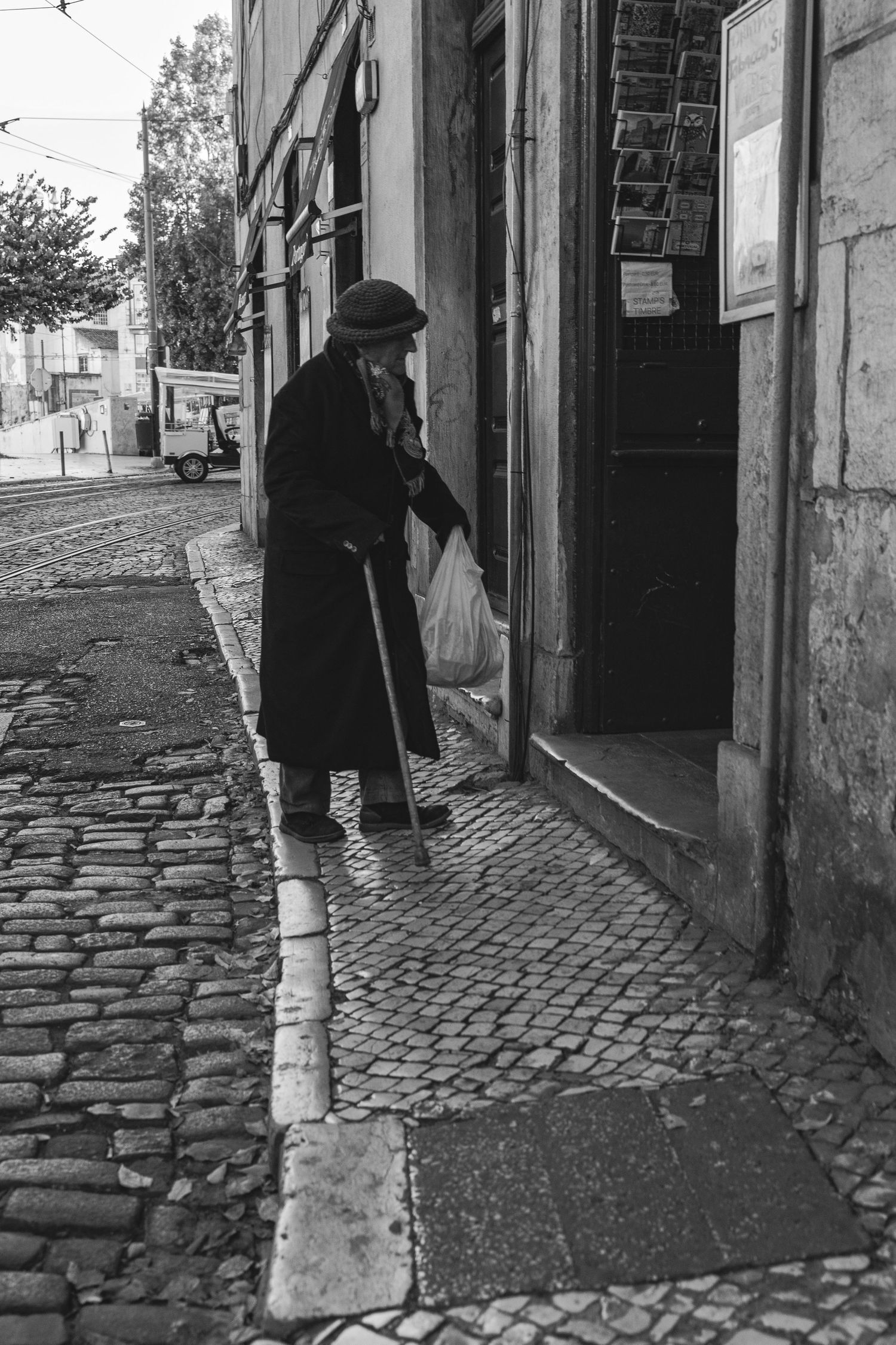 Black and white photo of elderly person with cane walking on cobblestone street in Lisbon.