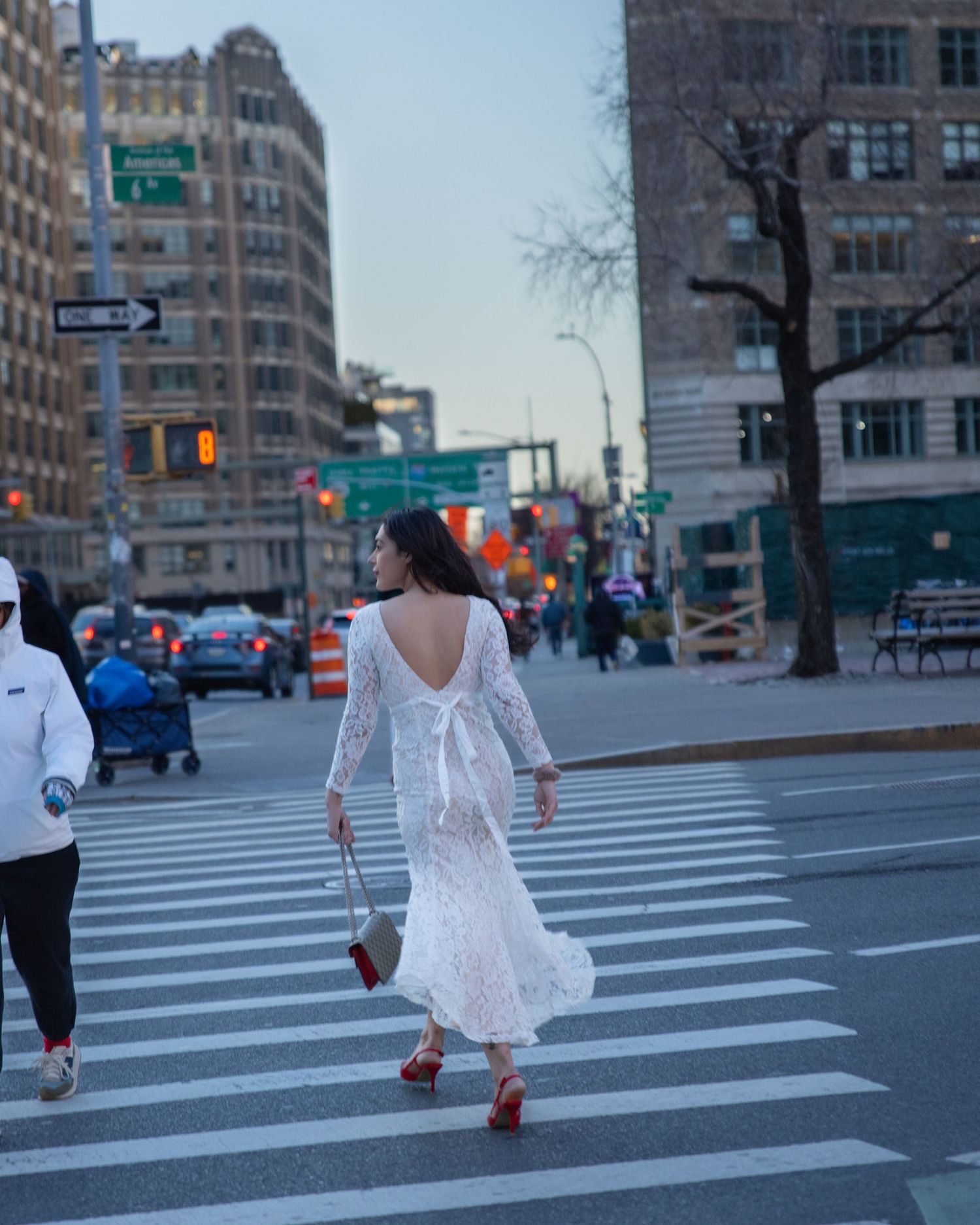 Woman in white lace backless dress and red heels crossing a busy New York City street intersection.