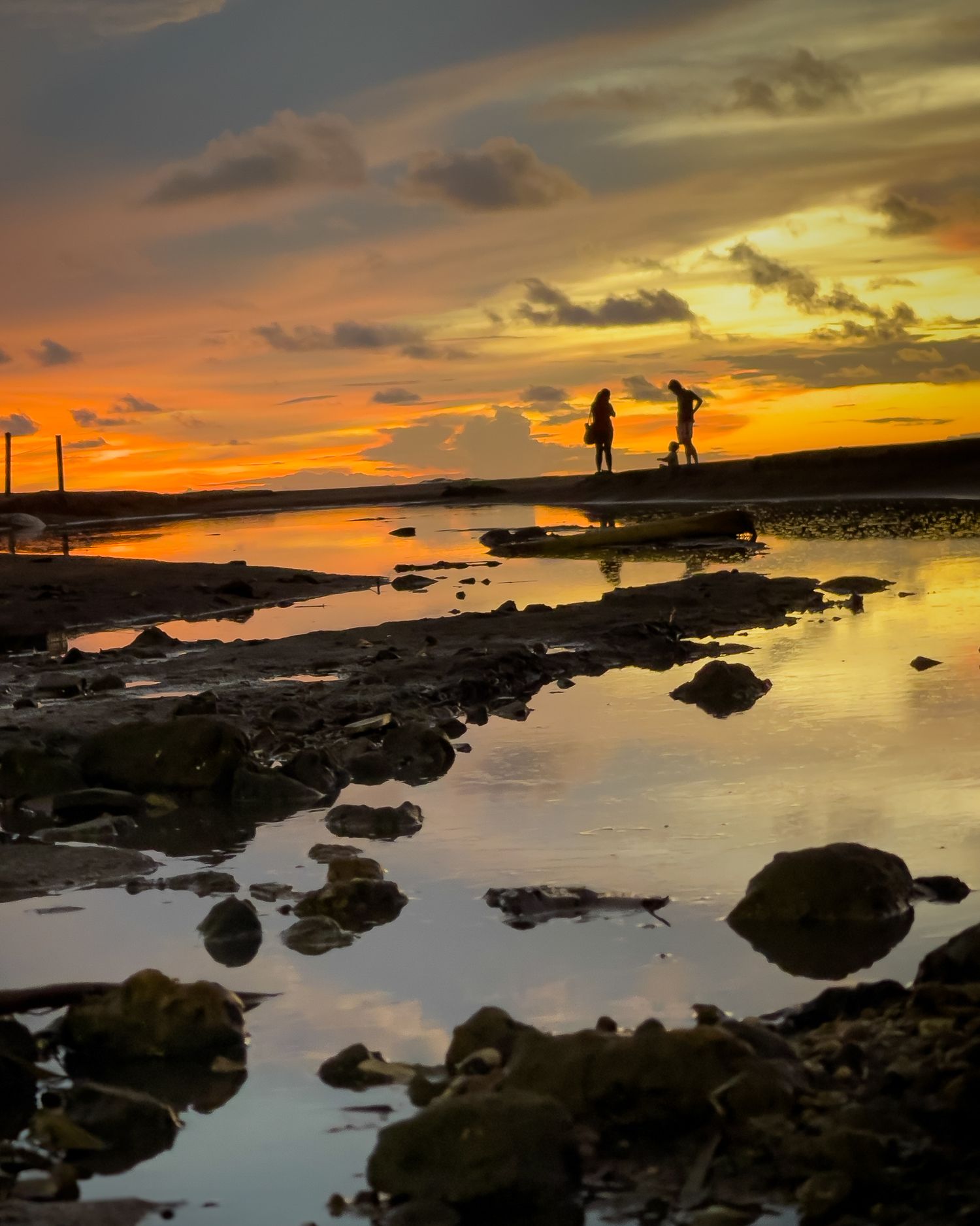 Silhouettes of people on rocky shoreline reflected in water during vibrant sunset.