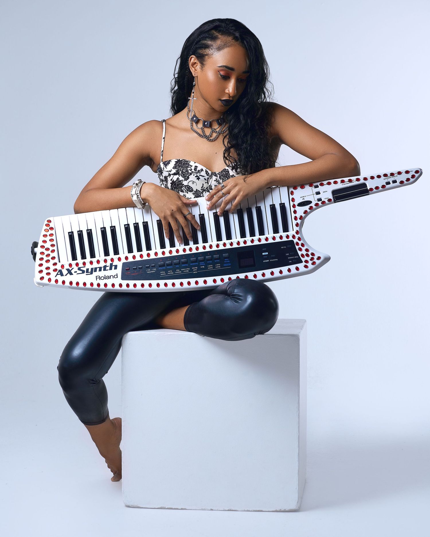 Young woman posing with a Roland AX-Synth keytar on a white cube in a bright studio setting.