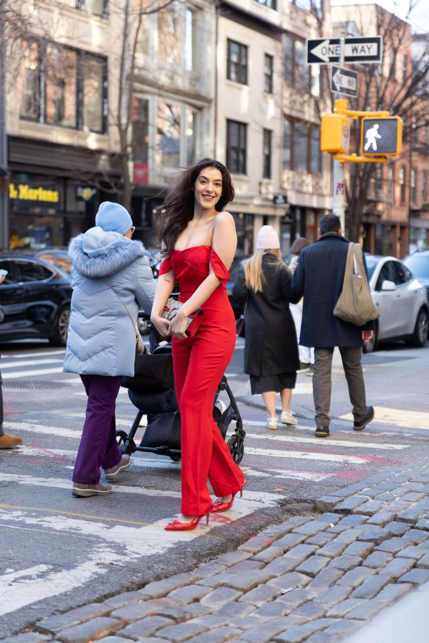 Woman in red off-shoulder jumpsuit and heels smiling on a New York City cobblestone street crosswalk.
