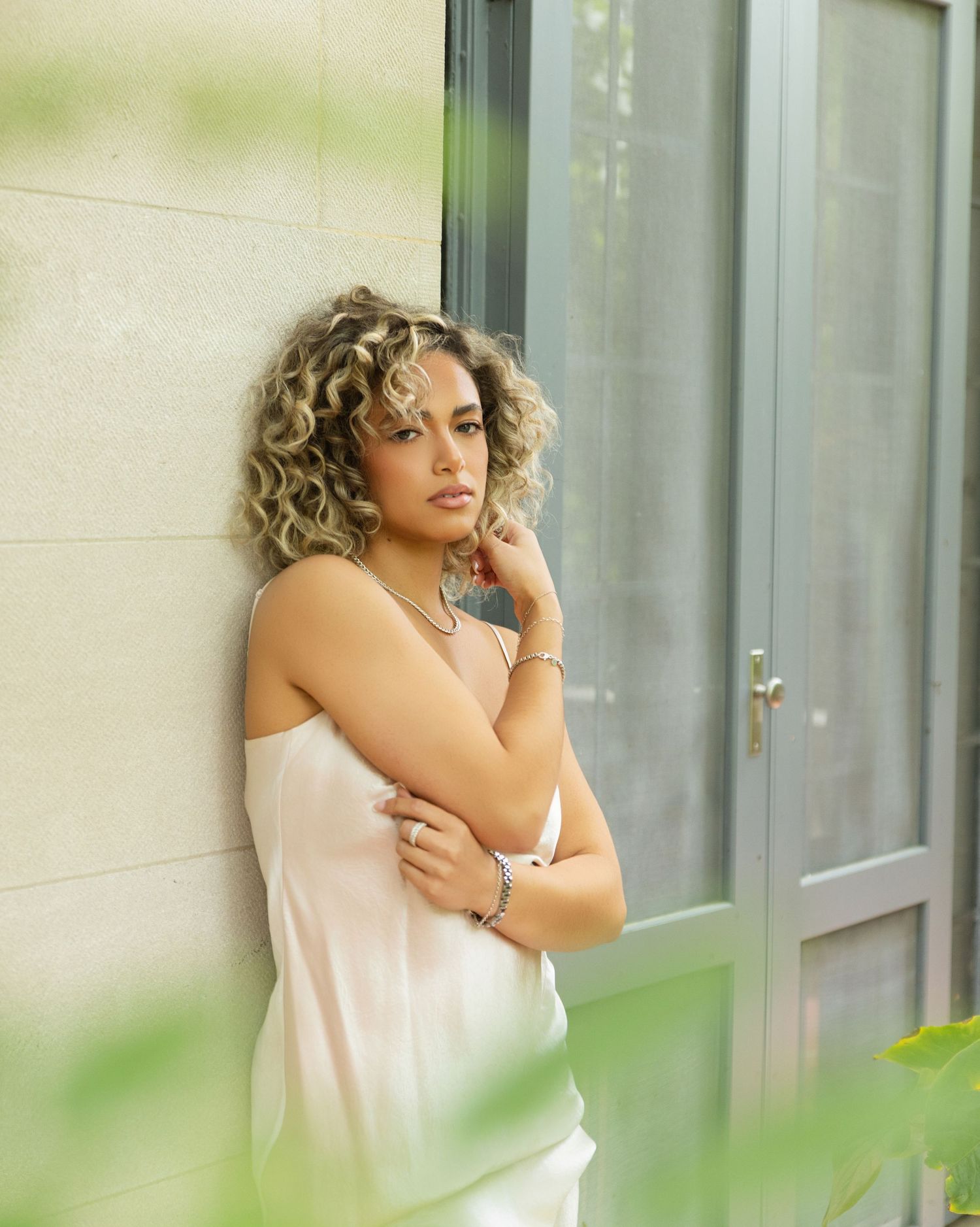 Woman with curly blonde hair in white satin slip dress leaning against a wall surrounded by soft greenery.