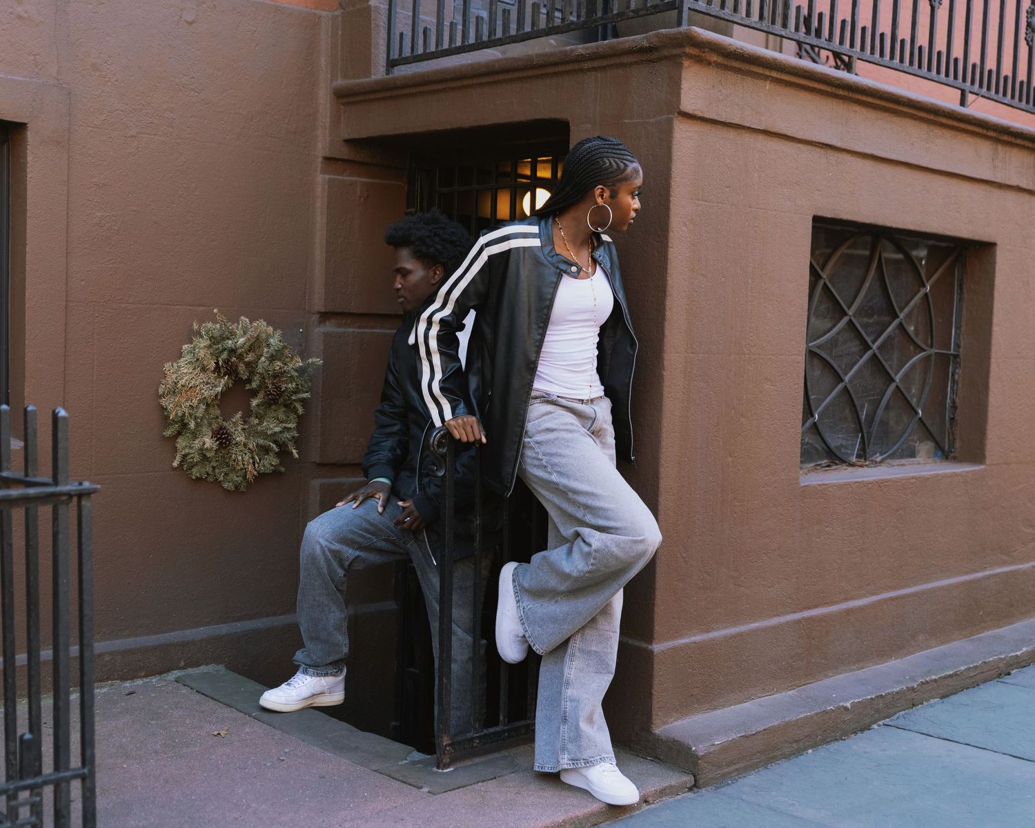 Two young people in casual streetwear leaning against a brownstone building entrance in an urban neighborhood.