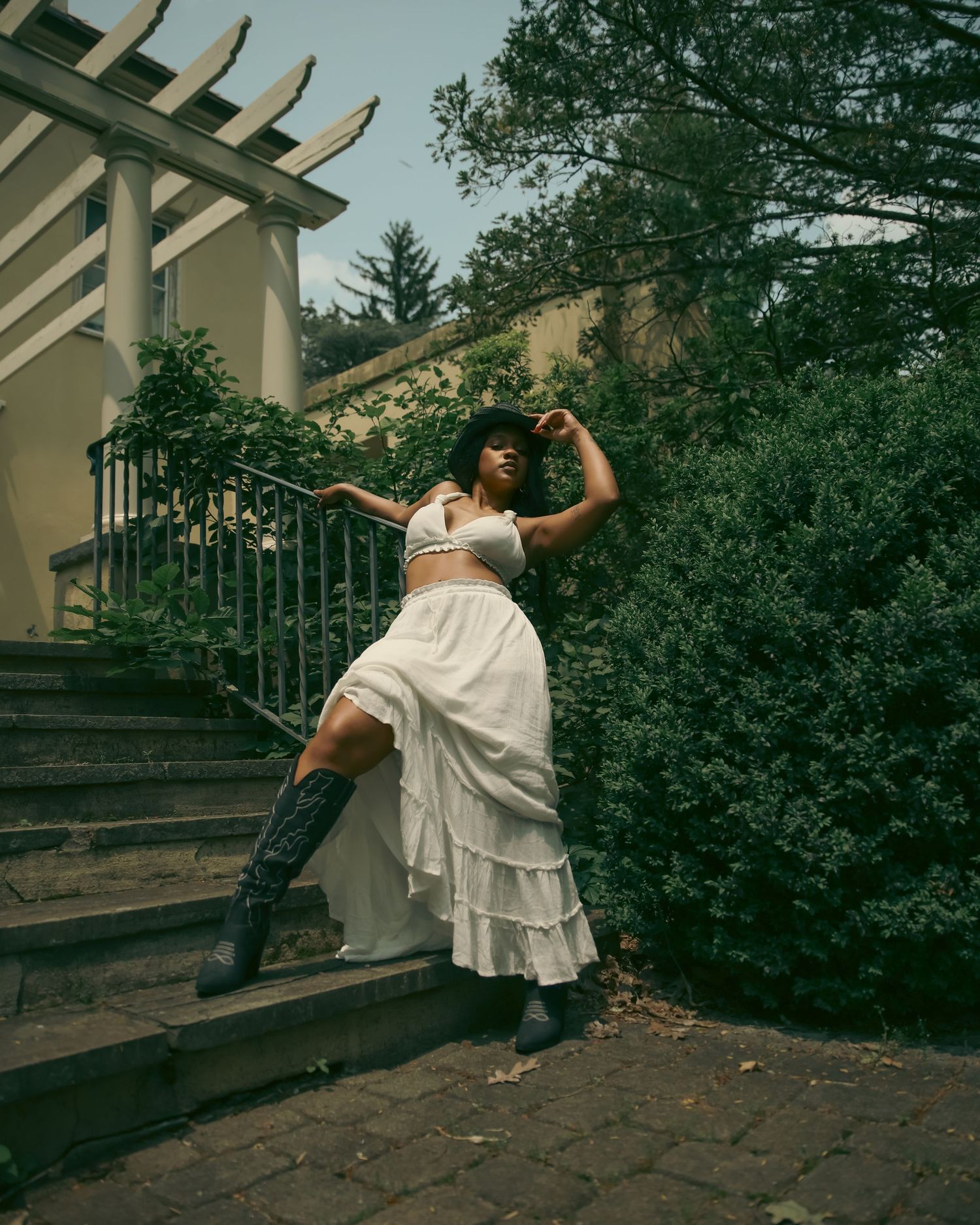Woman in white crop top and ruffled maxi skirt with cowboy boots posing on outdoor stone steps near greenery.