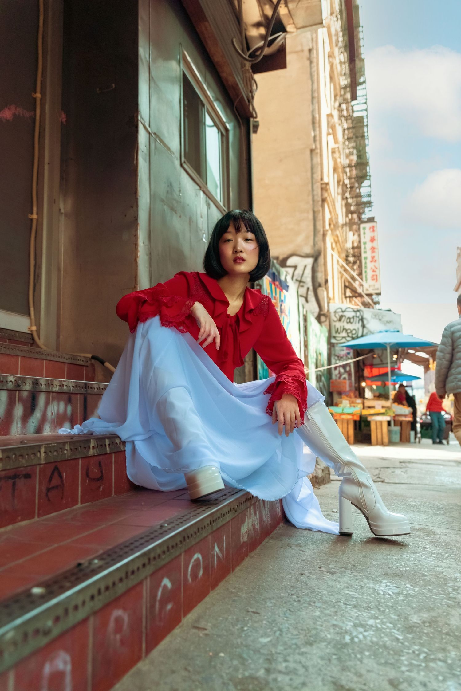 Young woman in red blouse and white skirt sitting on city steps in Chinatown urban environment.