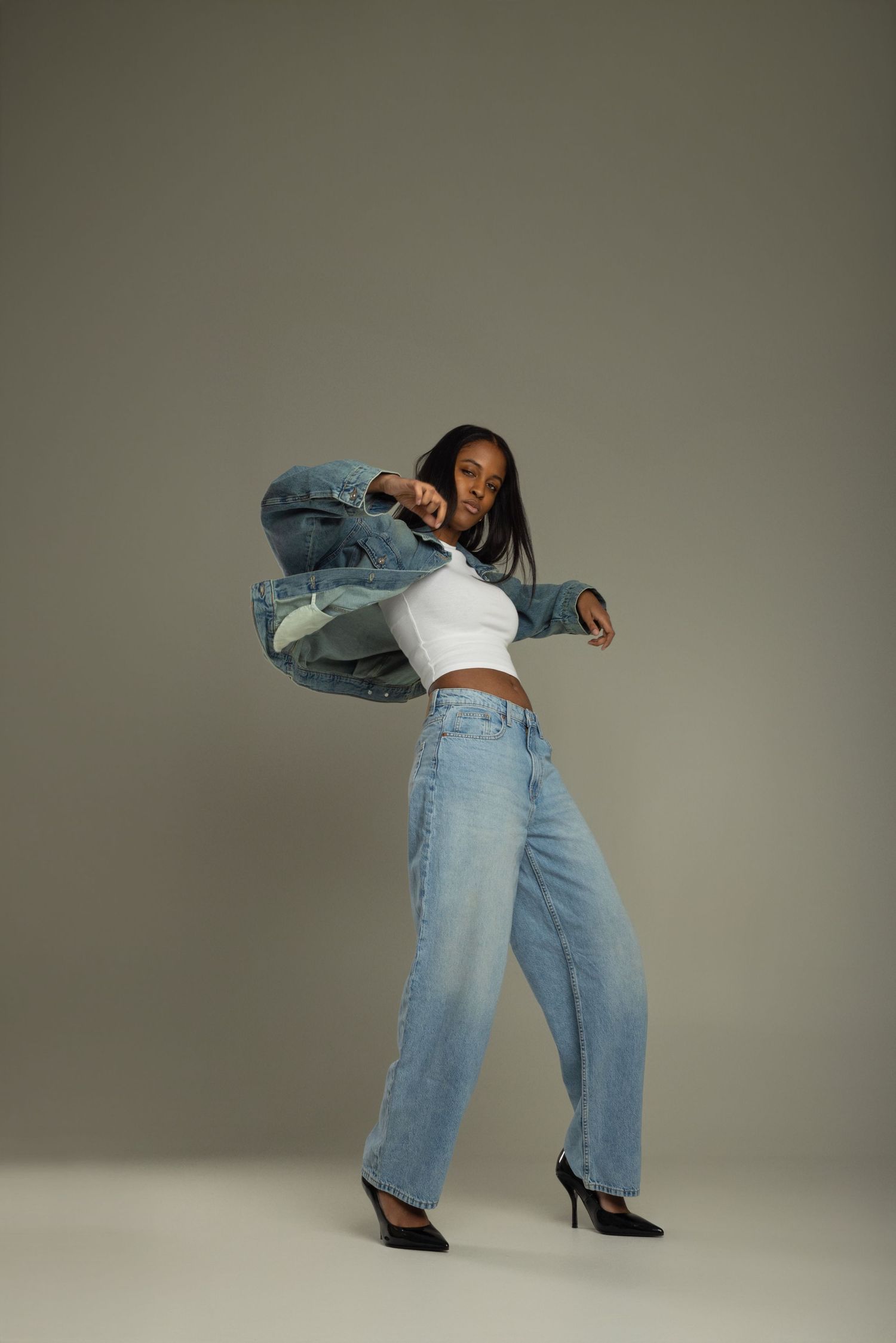 Young Black woman in denim jacket and wide-leg jeans dancing dynamically in studio on gray background.
